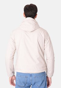 Giacca a cappuccio beige chiaro con una texture liscia e polsi elastici, abbinata a jeans in denim blu. Cuciture visibili e senza hardware prominente.