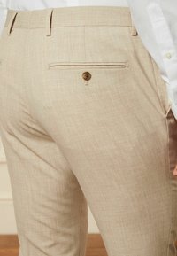Beige Hosen aus strukturiertem Stoff verfügen über eine Gesäßtasche mit einem Knopfverschluss. Das Design umfasst eine flache Front und einen schmalen Schnitt.