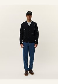 Cardigan negru cu închidere cu nasturi, două buzunare frontale și accente cu logo. Purtați peste o cămașă gri, cu blugi albaștri și pantofi maro. Design simplu.