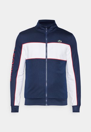Chaleco con cremallera en navy y blanco, con un alto cuello, que presenta acentos rojos y letras grandes en la manga. Fabricado con un tejido suave y ligero.