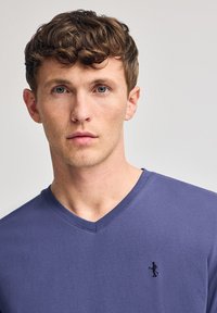 V-ringad t-shirt i solid mörklila bomull, med en liten broderad figur på vänster bröst. Slät textur och normal passform.