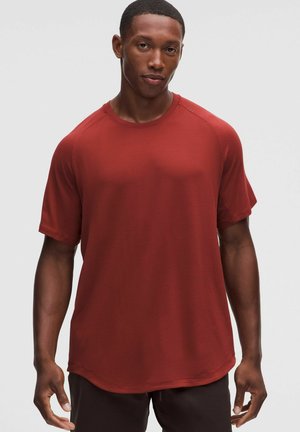 Mann trägt ein kurzärmeliges, rostfarbenes Sportshirt und dunkle Hose, steht vor einem einfarbigen hellen Hintergrund.