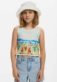 Svetlý bezrukávový crop top s kvetinovým vzorom v živých farbách, s volánovými okrajmi, na modrom pozadí krajiny, v kombinácii so svetlými džínovými šortkami.