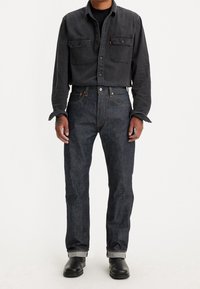 Dunkle Denim-Jeans mit engem Schnitt, Knopfleiste, traditionellem Fünf-Taschen-Design und kontrastierender Naht. Kombiniert mit einem schwarzen Hemd.