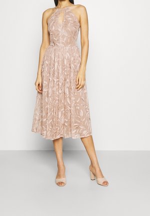 Frau trägt ein ärmelloses, knielanges, staubrosa Kleid mit floraler Spitzenüberlagerung und hautfarbene Sandalen mit Blockabsatz, steht auf einem hellen Boden.