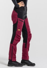 Maroon and black outdoor pants featuring a tapered design, zippered pockets, and reinforced ankle sections. Made of textured, durable fabric.