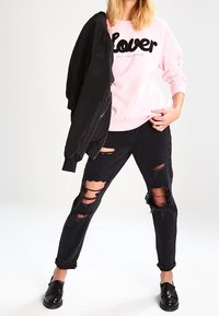 Roze sweatshirt met "Lover" in het zwart, gecombineerd met gescheurde zwarte jeans en zwarte loafers, aangevuld met een zwarte bomberjack.