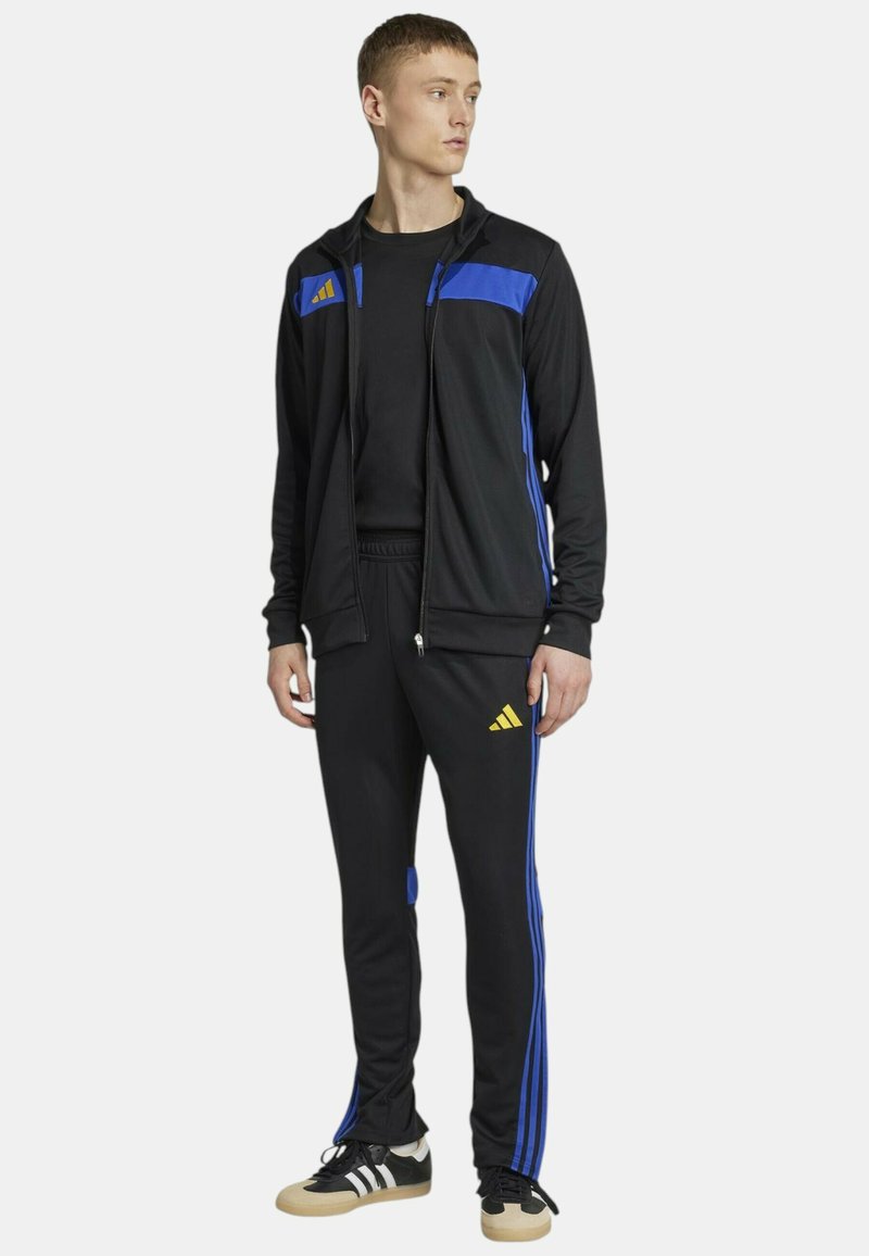 Giacca sportiva nera con accenti blu, chiusura frontale con cerniera e tre strisce sulle maniche. Abbinata a pantaloni neri coordinati e scarpe.