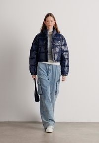 Tumed puffer-jakk, millel on läikiv viimistlus ja lühike disain. Hall pikkade varrukatega särk, helesinised laiad cargo-püksid ja valged spordijalatsid. Must kott.
