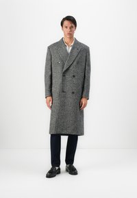 Junger Mann steht vor einem weißen Hintergrund und trägt einen langen grauen Zweireiher, dunkle Hosen und schwarze Fransenloafers.