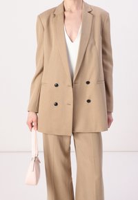 Beige dubbelzijdige blazer met zwarte knopen, gecombineerd met bijpassende broek. Het model houdt een kleine lichtroze handtas vast. Gladde stoftextuur.
