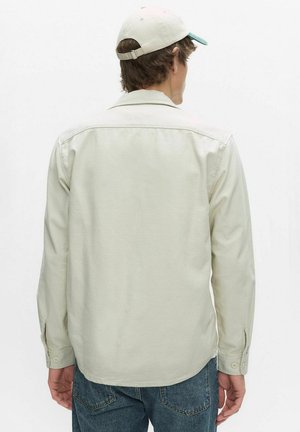 Mand iført lysebeige langærmet skjorte, blå jeans og en beige baseballkasket med ryggen vendt mod en ensfarvet hvid baggrund.