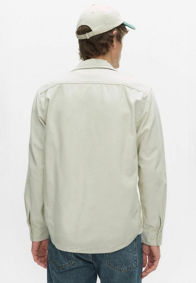 Man draagt een lichtbeige overhemd met lange mouwen, blauwe spijkerbroek en een beige baseballpet, kijkt weg tegen een effen witte achtergrond.