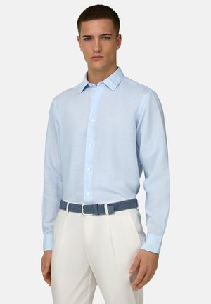 Jeune homme portant une chemise bleu clair à boutons, un pantalon blanc et une ceinture tressée bleue, debout devant un fond uni.
