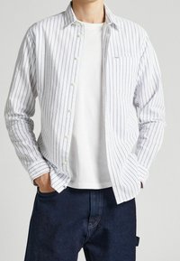 Camisa blanca de botones con rayas verticales azules, que cuenta con un bolsillo en el pecho y puños. Lleva puesta una camiseta blanca sencilla y pantalones azul oscuro.