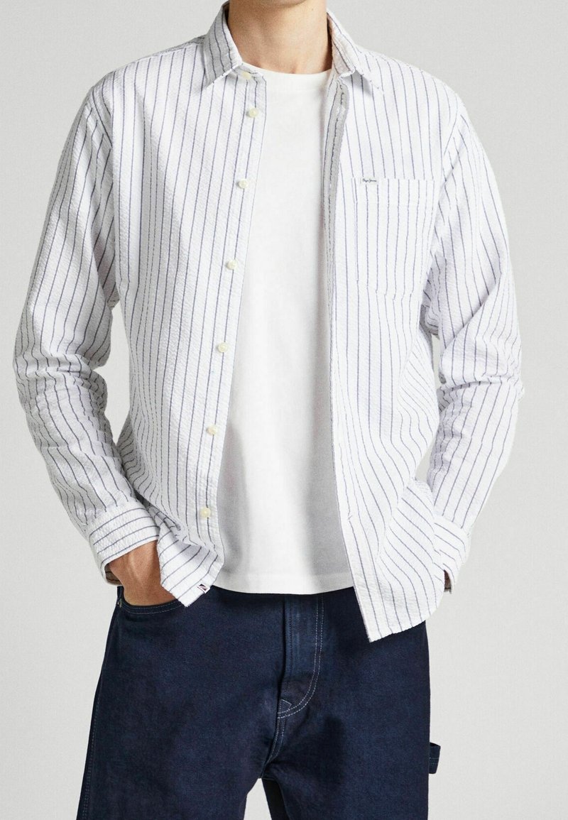 Camisa blanca de botones con rayas verticales azules, que cuenta con un bolsillo en el pecho y puños. Lleva puesta una camiseta blanca sencilla y pantalones azul oscuro.