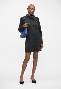 Abito grigio a maglia con colletto e maniche lunghe, abbinato a una piccola borsa intrecciata blu e scarpe con tacco a punta nere. Texture liscia.