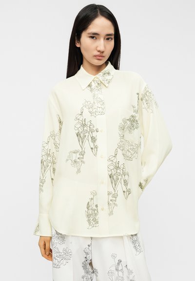 Μπλούζα σε κρεμ χρώμα από ελαφρύ ύφασμα με μαύρα σχέδια λουλουδιών σε γραμμή. Διαθέτει κλασικό γιακά και κλείσιμο με κουμπιά μπροστά.