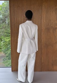 Blazer texturé blanc et pantalon large, avec une taille ceinturée et un motif floral subtil, associés à des chaussures à talons hauts argentées.