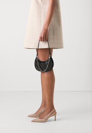 Nainen beigenvärisessä lyhyessä mekossa ja nudensävyisissä slingback-koroissa, joka pitää mustaa niitillä koristeltua käsilaukkua kahvasta yksiväristä taustaa vasten.