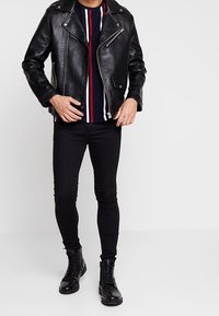 Černá kožená motorkářská bunda se stříbrnými zipy, nošená přes pruhované tričko v námořnické modři, červené a bílé, v kombinaci s černými úzkými džíny a botami.