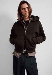 Junge Person mit schulterlangem Haar, trägt einen schwarzen Kapuzenpullover mit Reißverschluss über einem gestreiften Hemd und dunkler Hose, Hände in den Taschen des Kapuzenpullovers.