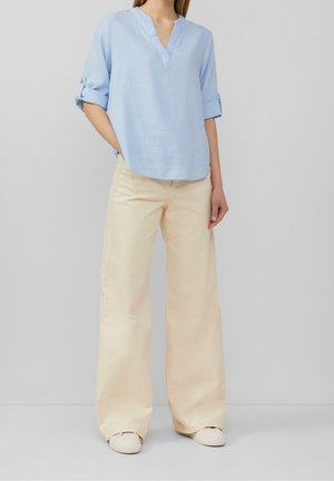 Personne portant un chemisier bleu clair à col en V avec manches retroussées, un pantalon large crème et des chaussures blanches sur un fond gris uni.