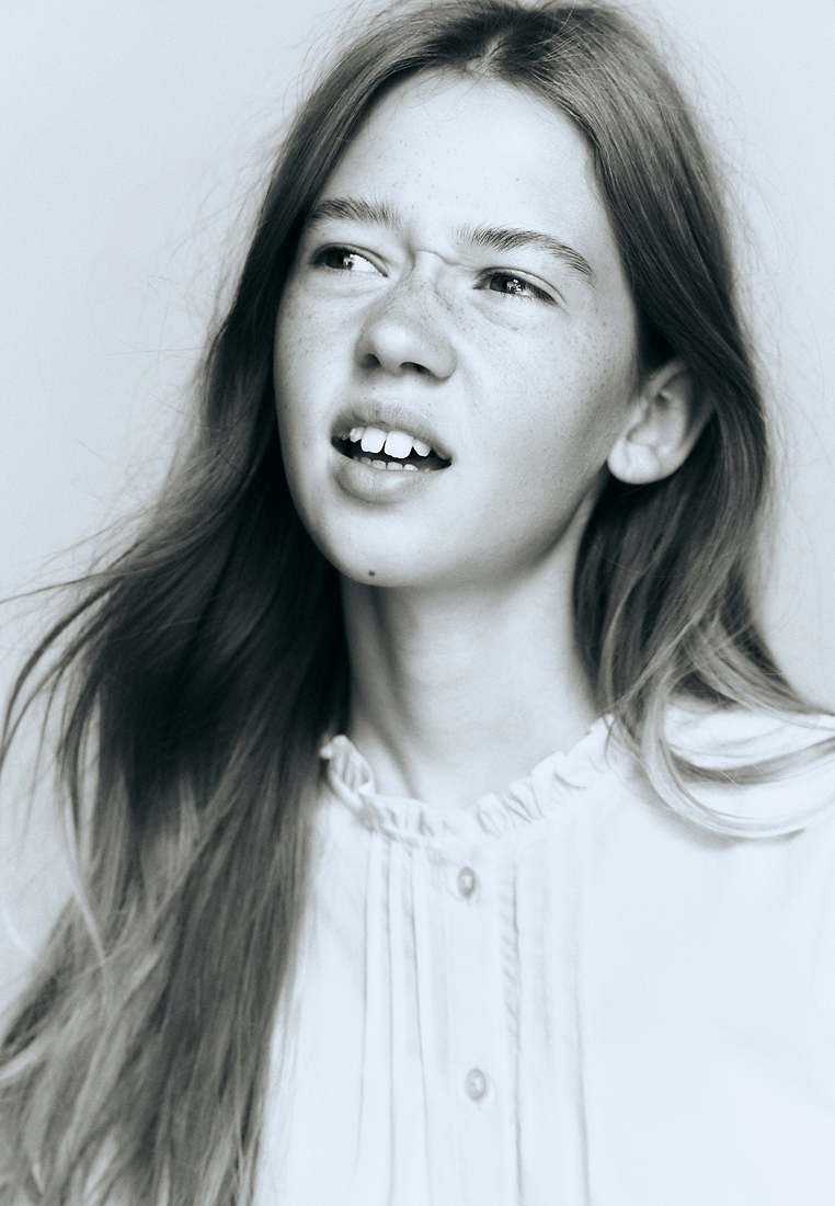 Jeune fille aux longs cheveux et aux taches de rousseur regarde sur le côté avec une expression perplexe, portant un chemisier blanc à boutons.