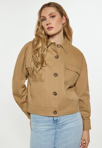 Beige verkürzte Jacke aus Baumwolle mit Hemdkragen, zwei vorderen Taschen und braunen Knöpfen, kombiniert mit einem hellblauen Jeansrock.