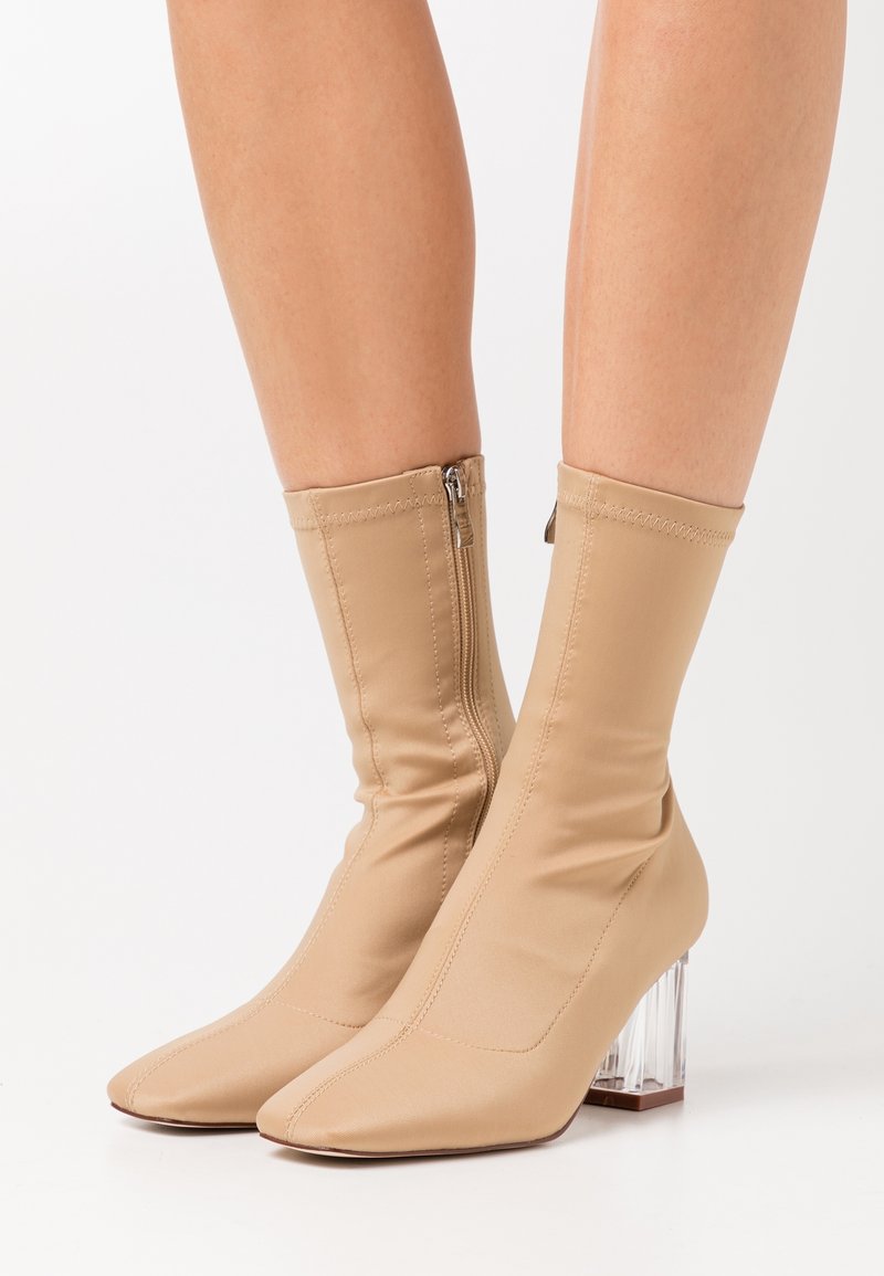 Botines tipo calcetín en beige con tacón bloque transparente, punta puntiaguda y cierre de cremallera, con una textura de tela suave y elástica.