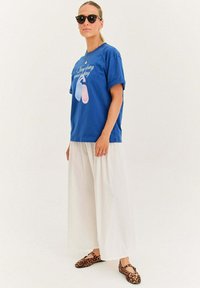 Modrá bavlnená tričko s krátkymi rukávmi, ktoré má grafický potlač, biele široké nohavice a hnedé topánky s leopardím vzorom.