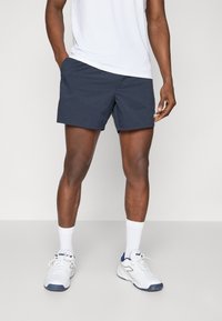 Marineblå treningsshorts med en glatt tekstur, med sidelommer og en logo-detalj. Sammen med hvite sokker og sporty sko.