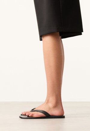 Seitenansicht einer Person, die schwarze flache Zehensandalen mit schwarzen, oberhalb des Knöchels gekürzten Hosen trägt und auf einem hellen Boden steht.