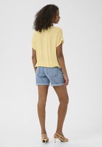 Haut jaune à manches courtes avec une coupe décontractée, associé à un short en denims retroussé. La modèle porte des sandales à talons jaunes. Texture lisse et style décontracté.