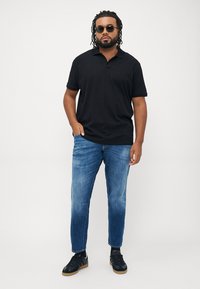 Crna polo majica kratkih rukava, plave traperice, crne patike. Model ima labavu kovrčavu kosu i nosi sunčane naočale. Neutralna pozadina.