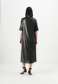 Donna con capelli scuri di lunghezza spalla che indossa un vestito midi nero lucido e sandali neri con tacco alto e cinturini alla caviglia, girata di spalle.