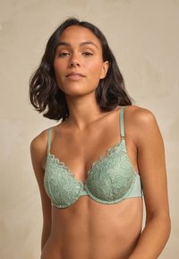 Žalios mėtų spalvos liemenėlė su nėrinių kaušeliais su banguotais kraštais, reguliuojamomis petnešėlėmis ir lygiu apačiu. Matomi auksinės spalvos metaliniai akcentai.