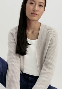 Lichtgrijze, gebreide cardigan met ribbelstructuur, open voorkant en lange mouwen, gecombineerd met een witte ronde hals t-shirt en donkerblauwe jeans.