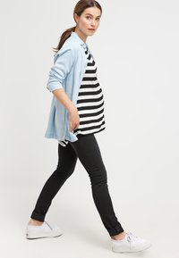 Top de maternidade preto e branco às riscas, sobreposto a uma camisa de chambray azul claro, combinado com jeans skinny pretos e sapatilhas brancas.