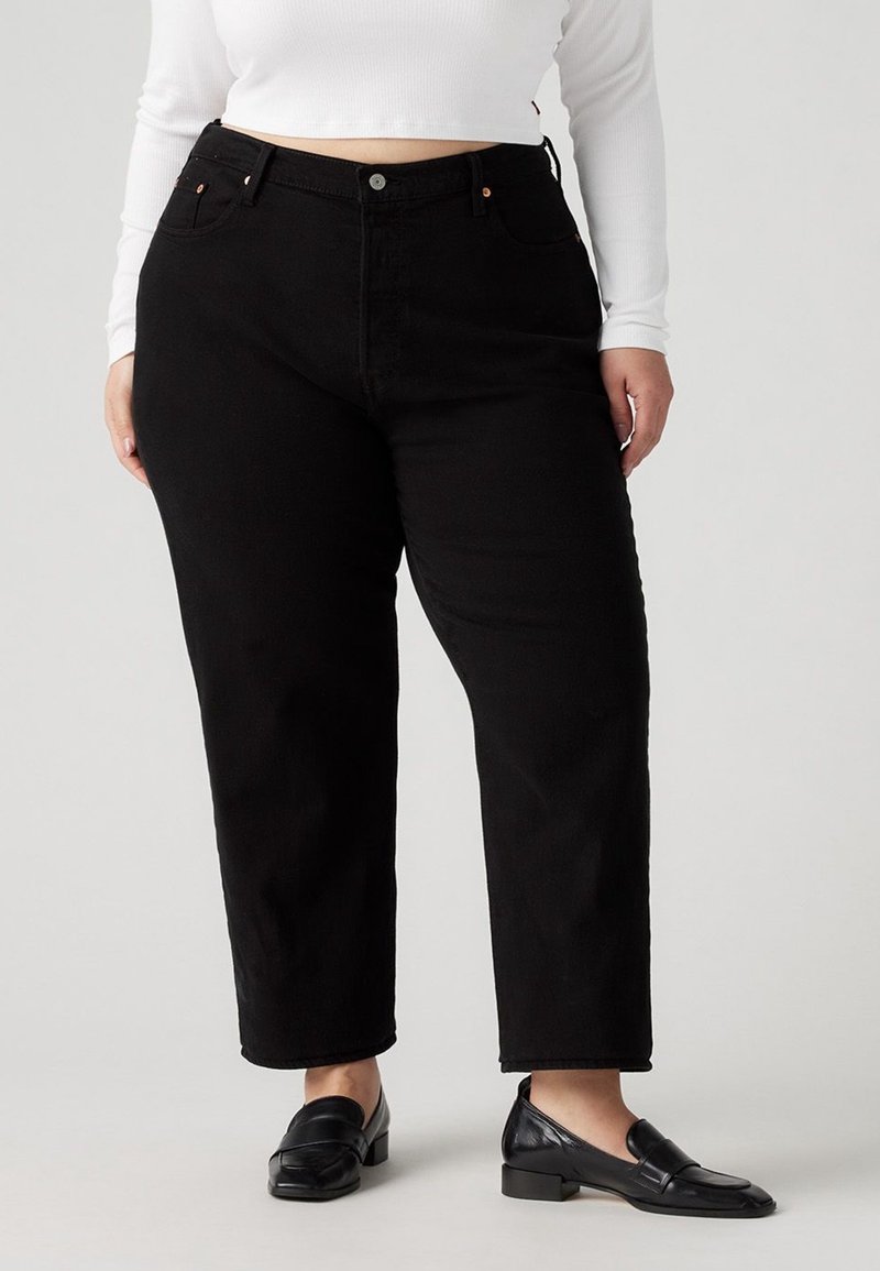 Personne aux formes portant un jean noir taille haute, un top blanc à manches longues et court, et des mocassins en cuir noirs, debout sur un fond uni.