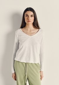 Mlada ženska z dolgimi rjavimi lasmi, oblečena v belo majico z V-izrezom in dolgimi rokavi ter svetlo zelene hlače s vezalko, stoji pred enobarvnim ozadjem.