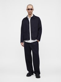 Homme portant des lunettes de soleil foncées, une veste et un pantalon noirs, une chemise blanche, et des chaussures noires épaisses, debout avec une main dans la poche.