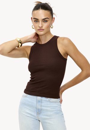 Jeune femme portant un haut sans manches marron et un jean bleu clair, accessoirisée avec des boucles d'oreilles en or et un bracelet, posant avec la main derrière le cou.