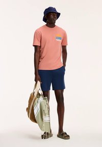 Pink cotton t-shirt with a graphic pocket, navy shorts, green beach towel, woven bag, and navy bucket hat, worn with green sandals.