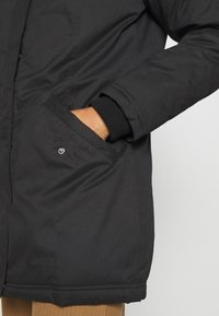 Black insulated jacket with a smooth matte finish, featuring ribbed cuffs, front pocket, and a straight cut design.