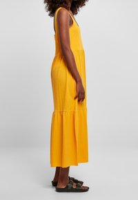 Żółta bawełniana sukienka maxi o bezrękawowym kroju, dopasowanej górze i falbaniastej spódnicy. Noszona z ciemnymi sandałami. Prosta, gładka tekstura.