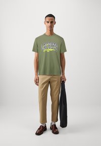 Olīvu zaļa T-kreklu ar dzeltenu un zilu "LACOSTE" grafiku, apvienota ar bēšiem biksēm un brūnām loafers, tur melnu jaku.