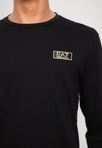 Černá dlouhá rukávová košile vyrobená z bavlny s kulatým výstřihem, která má na hrudi zlatou výšivku loga EA7 Emporio Armani.
