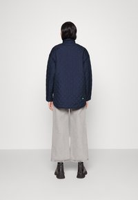 Chaqueta acolchada de marina con un alto cuello, que presenta un dobladillo redondeado y un pequeño acento verde. Combinada con pantalones de mezclilla gris ancho y botas negras.