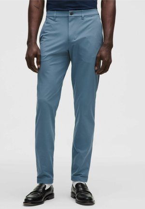 Mand iført smalle lyseblå bukser, sorte læderloafers og hvide sokker, stående foran en ensfarvet baggrund.